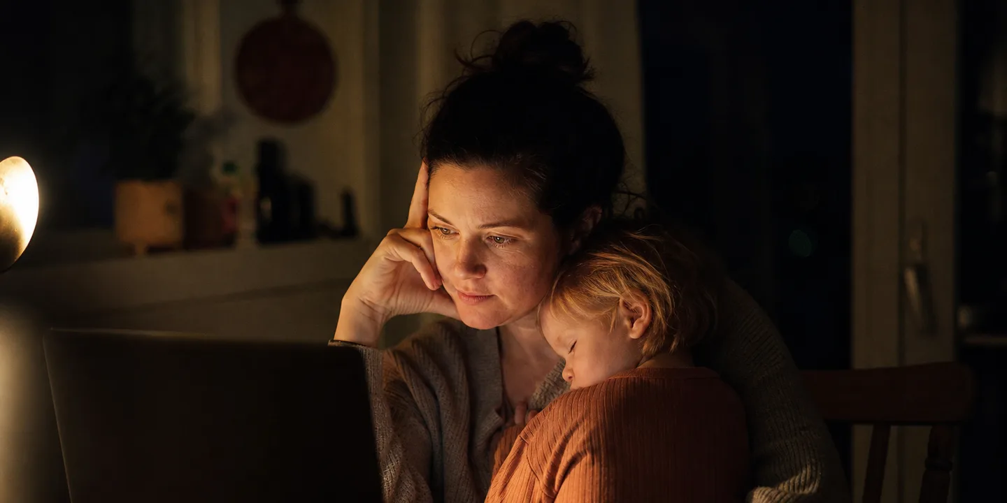 A mother sitting at a dark kitchen table late at night, lit only by her laptop and a small lamp, with a sleeping toddler resting against her shoulder