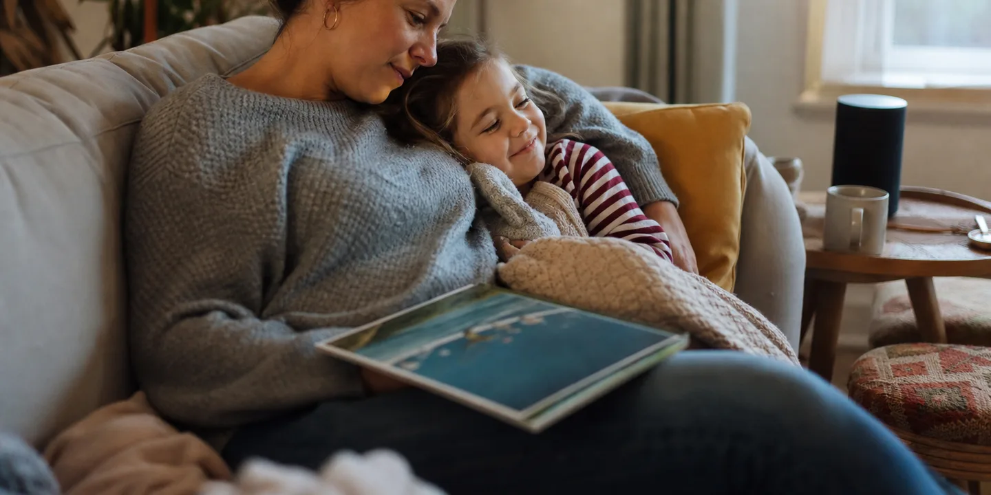 A mother and her young daughter cuddled under blankets on a sofa in the late afternoon, listening to a story playing from a small speaker on the side table, a square picture book resting on the mother's lap